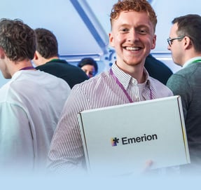 Emerion-Mitarbeiter mit Firmenpaket im Arm, im Hintergrund Kolleginnen und Kollegen (AI generated)