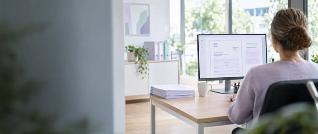 Mitarbeiterin im Büro prüft am Bildschirm ein digitales Dokument, neben dem Monitor liegt ein Stapel Papierbelege (KI-generiert)