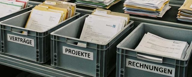 Graue Sortierkisten mit Papierakten, beschriftet mit den Kategorien Verträge, Projekte und Rechnungen.